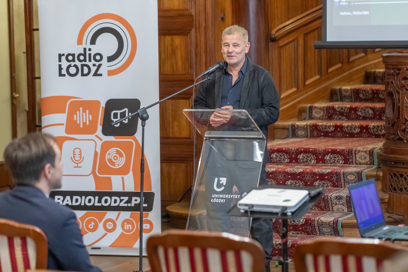 Na zdjęciu widać mężczyznę stojącego za mównicą i mówiącego coś do ludzi siedzących na sali.