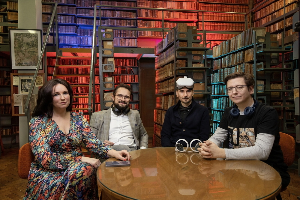 Cztery osoby siedzące przy stole, patrzą w stronę obiektywu. Otaczają je regały książek. Pomieszczenie wypełnia kolorowa poświata. Każda z osób ma przy sobie jakieś urządzenie elektroniczne: słuchawki, telefon lub okulary do projekcji wirtualnej.