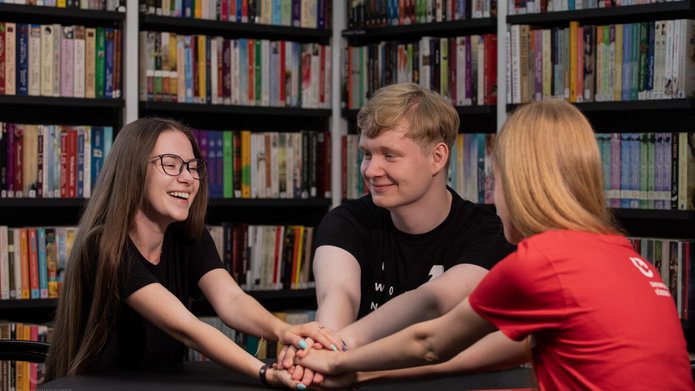 2 studentki i student siedzą w bibliotece przy regałach z książkami i trzymają się za ręce 