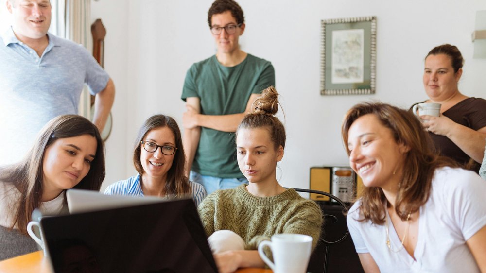 group of smiling young people sitting in a room, looking at a laptop screen