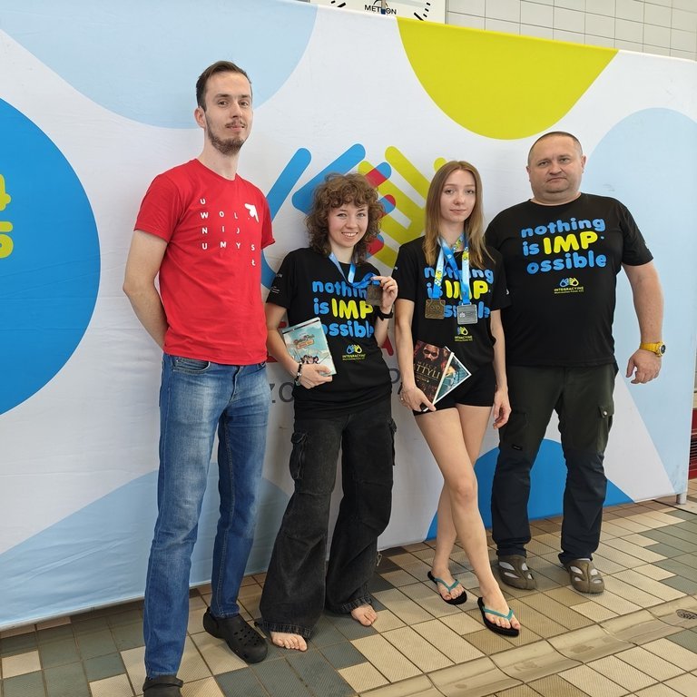 Cztery osoby pozują na tle kolorowego banera z napisem „sezon 2024/2025”. Dwie młode kobiety stoją po środku, ubrane w czarne koszulki z napisem „nothing is IMPossible”; trzymają medale i książki. Towarzyszą im dwaj mężczyźni – jeden w czerwonej koszulce po lewej i drugi w czarnej po prawej. Wszyscy stoją na basenowej posadzce, sugerując udział w wydarzeniu sportowym lub konkursie.