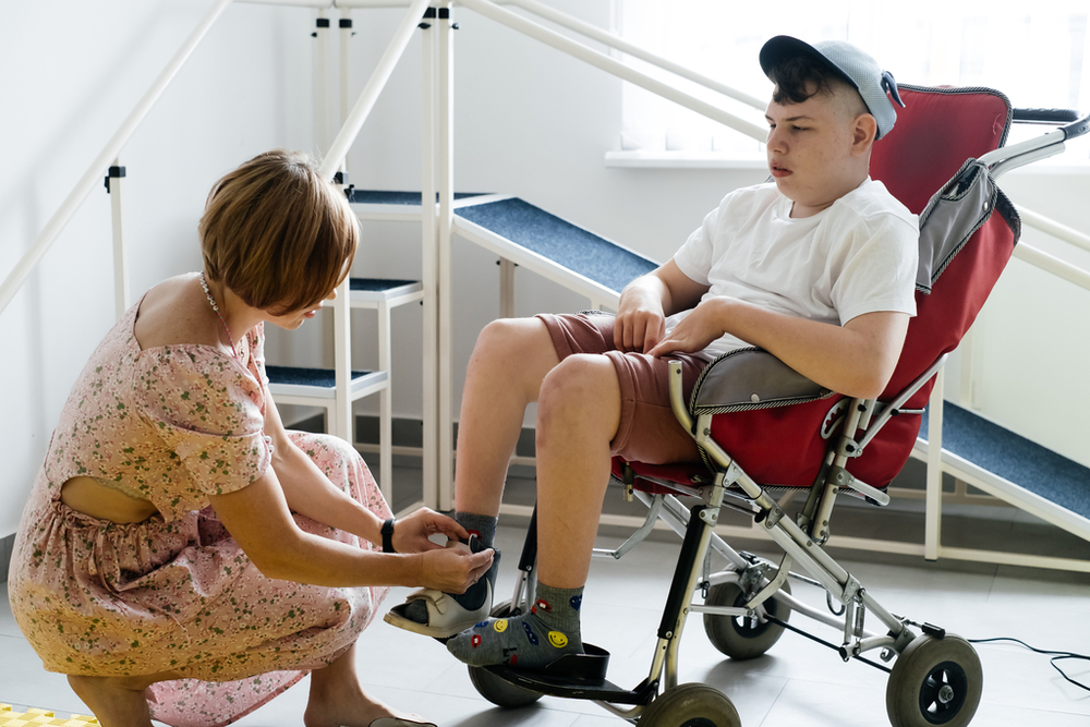 A mother helps a 17-year-old disabled boy with cerebral palsy in a wheelchair put on his shoes before going for a walk.