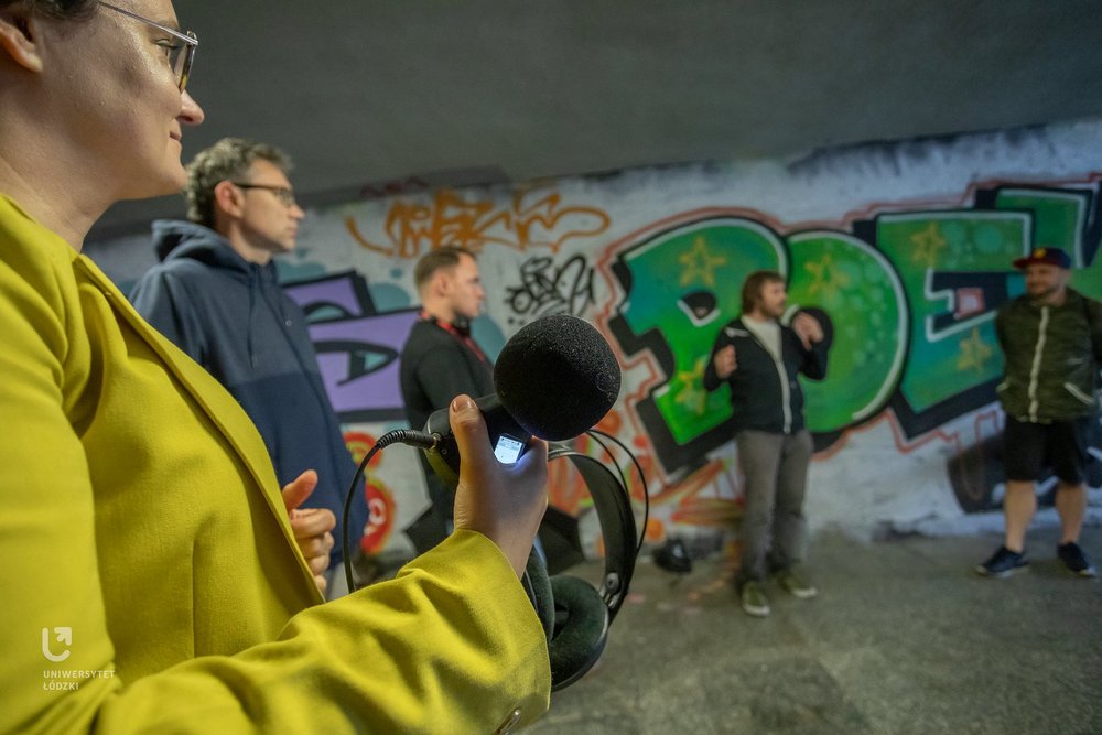 Na zdjęciu widać grupę ludzi. Stoją przejściu podziemnym. Trzymają w rękach urządzenia do nagrywania dźwięków. 